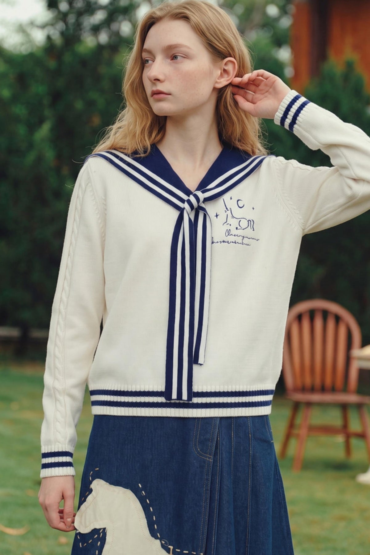 A woman wearing a cream and navy blue knit sweater with a sailor collar and tie, paired with a denim skirt, standing outdoors in a garden setting.