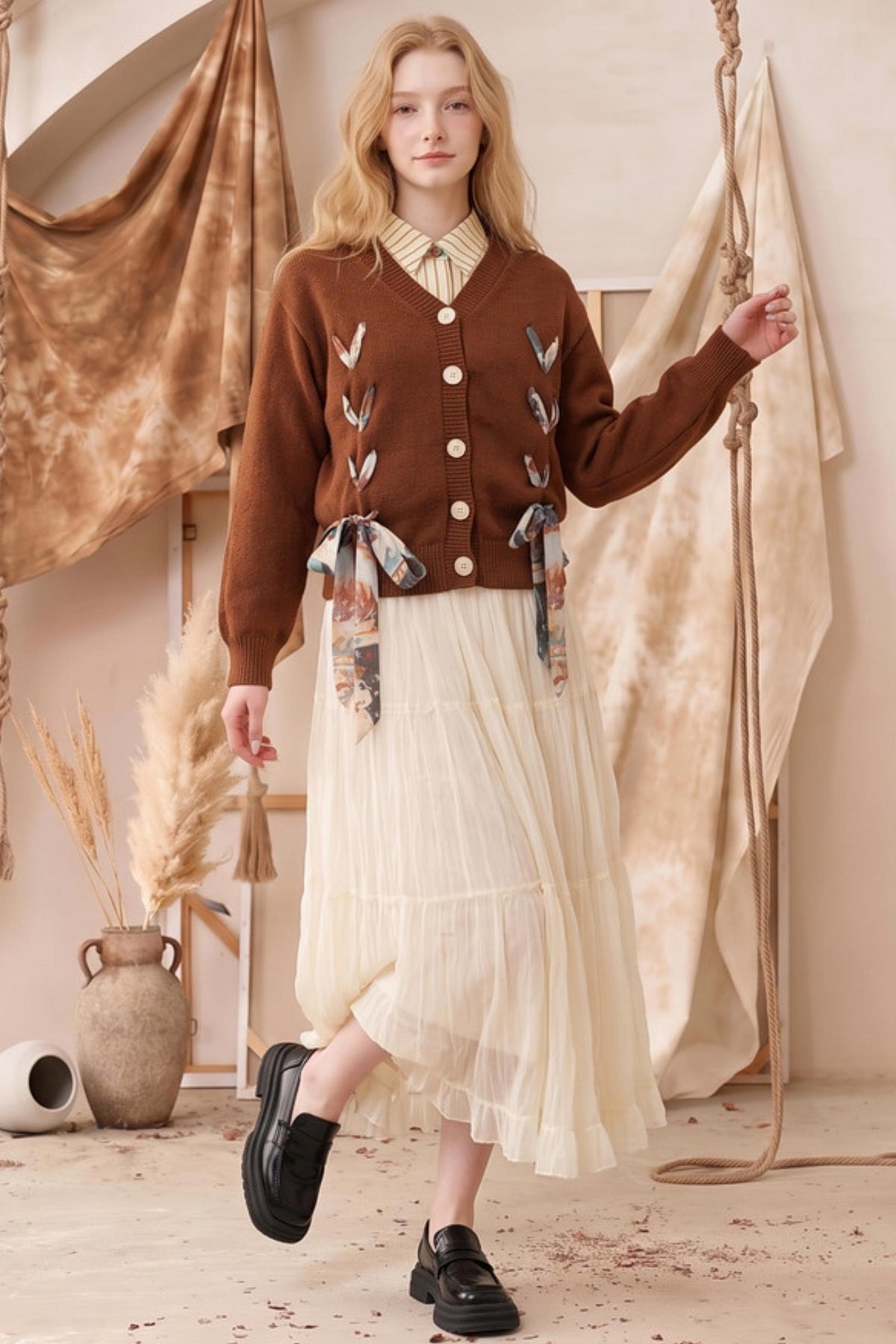 A woman models a brown button-up cardigan with embroidered details and patterned ribbon ties, paired with a cream pleated midi skirt and black loafers, set against a warm bohemian backdrop with hanging fabrics and dried pampas grass.