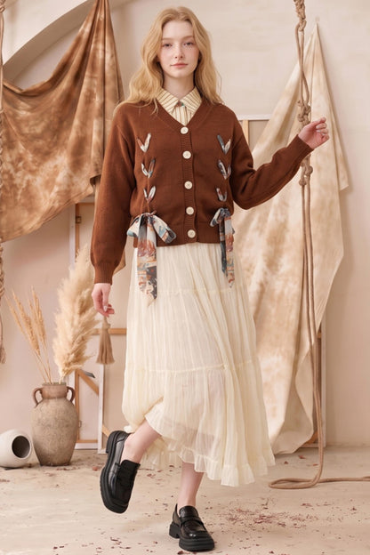 A woman models a brown button-up cardigan with embroidered details and patterned ribbon ties, paired with a cream pleated midi skirt and black loafers, set against a warm bohemian backdrop with hanging fabrics and dried pampas grass.