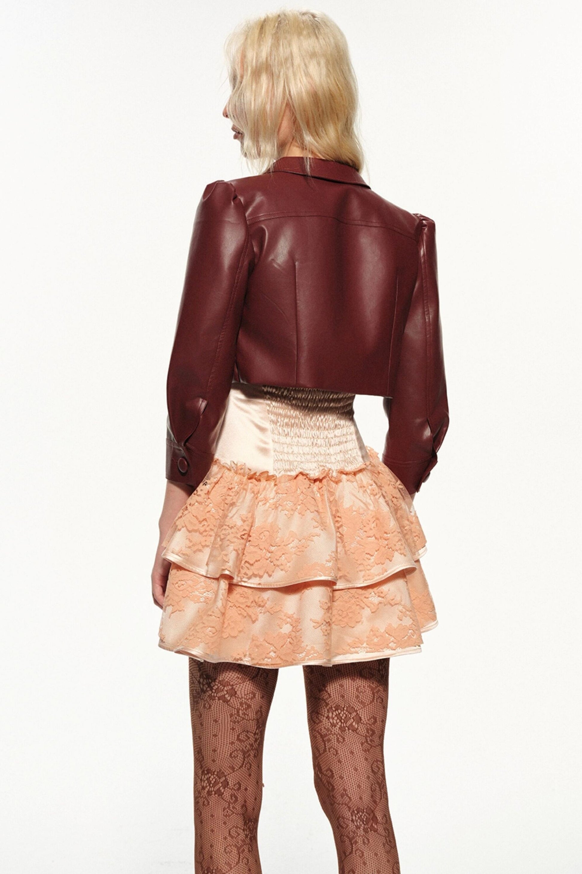 A model shown from the back wearing a dark burgundy faux-leather cropped jacket, a peach and cream ruffled lace mini skirt, and sheer patterned tights against a plain white background.
