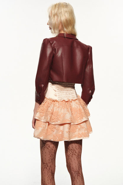 A model shown from the back wearing a dark burgundy faux-leather cropped jacket, a peach and cream ruffled lace mini skirt, and sheer patterned tights against a plain white background.