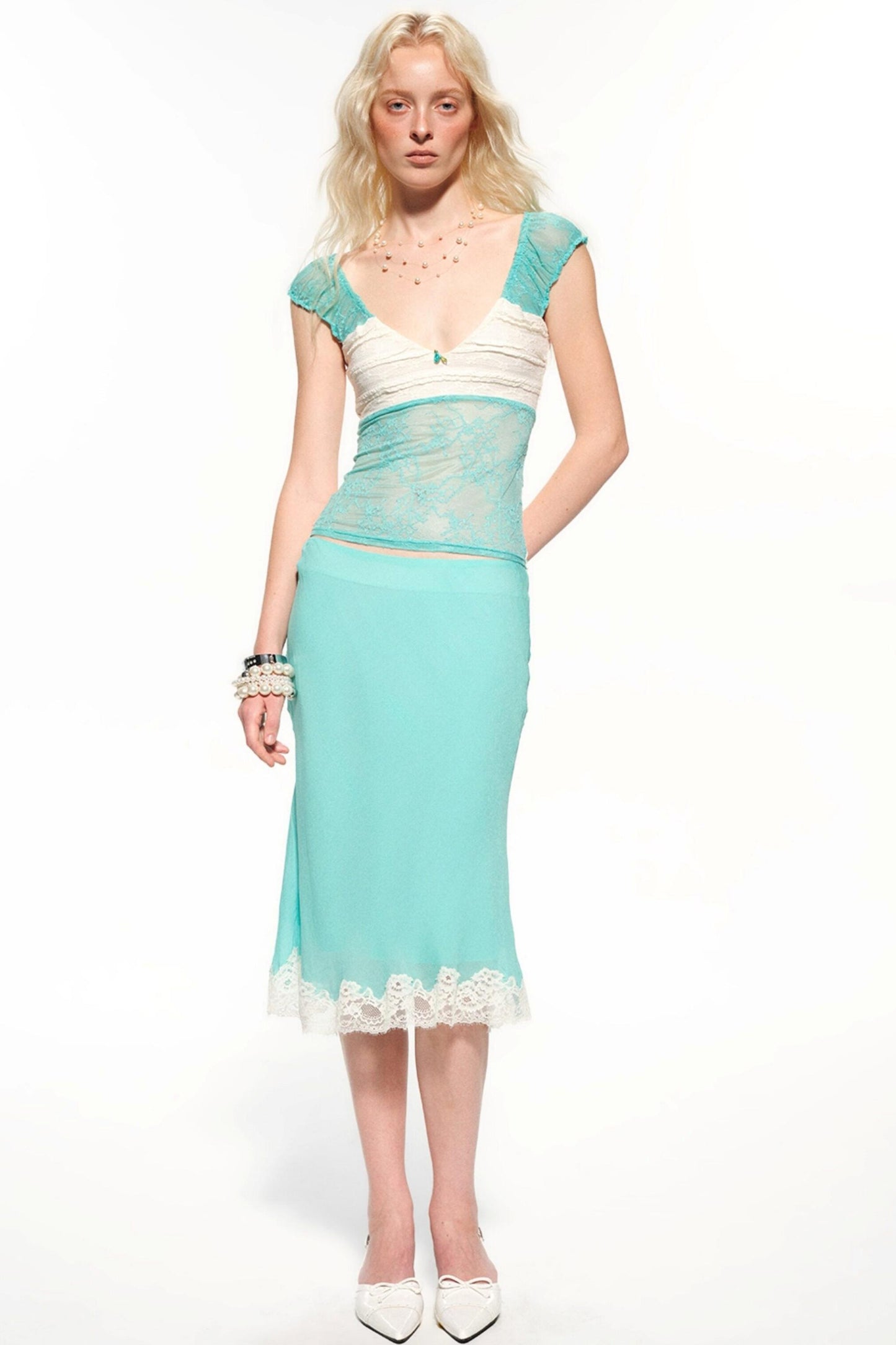 A blonde model wearing a two-piece dress with a turquoise and white color-blocked bodice featuring lace applique, and a matching solid turquoise skirt with a white lace hem, paired with white pointed-toe heels.