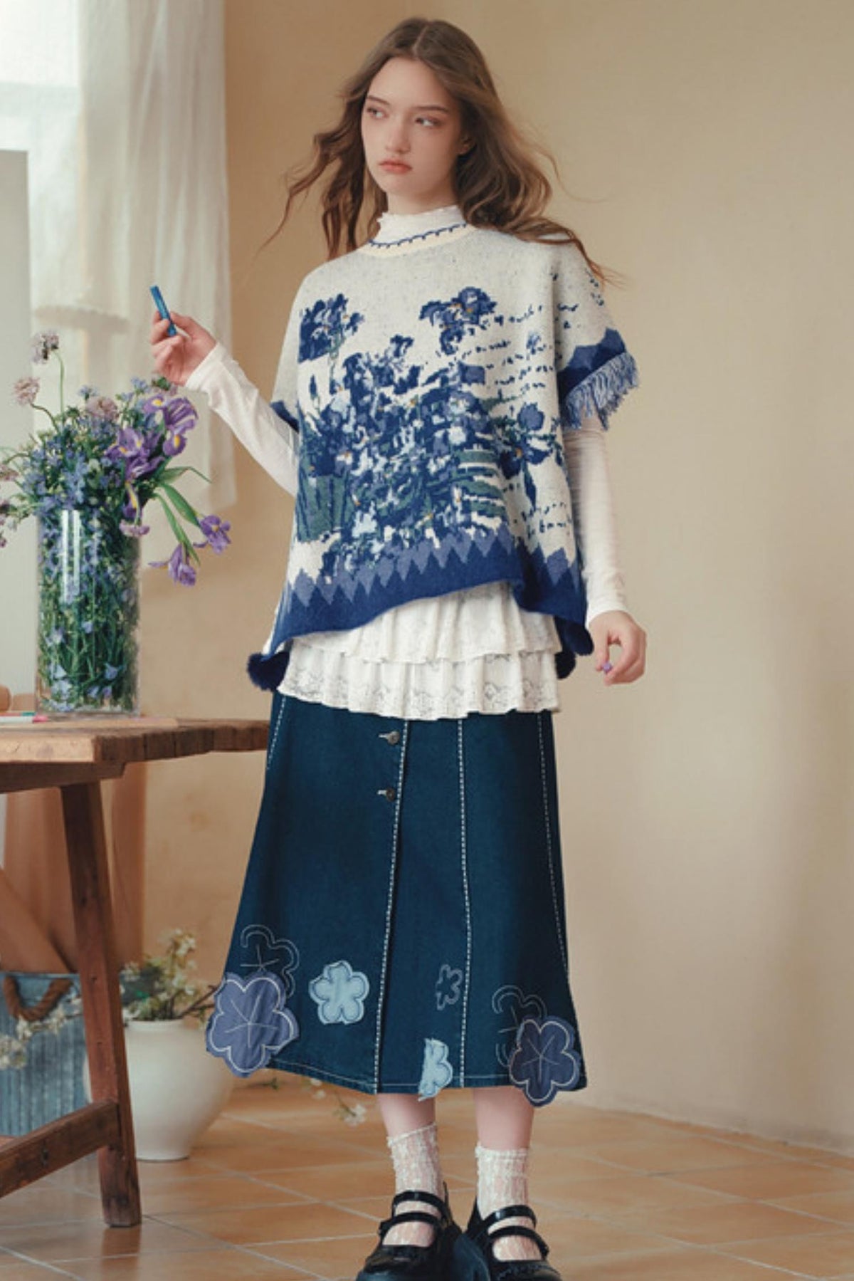 A woman stands indoors wearing a cream knit poncho-style sweater with blue floral and geometric patterns over a white ruffled long-sleeve top, paired with a dark blue denim skirt featuring light blue floral embroidery near the hem. She is holding a small blue object in one hand, with a vase of flowers and a wooden table visible in a softly lit room.