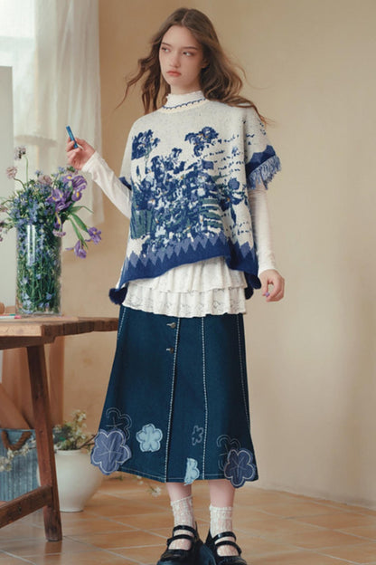 A woman stands indoors wearing a cream knit poncho-style sweater with blue floral and geometric patterns over a white ruffled long-sleeve top, paired with a dark blue denim skirt featuring light blue floral embroidery near the hem. She is holding a small blue object in one hand, with a vase of flowers and a wooden table visible in a softly lit room.