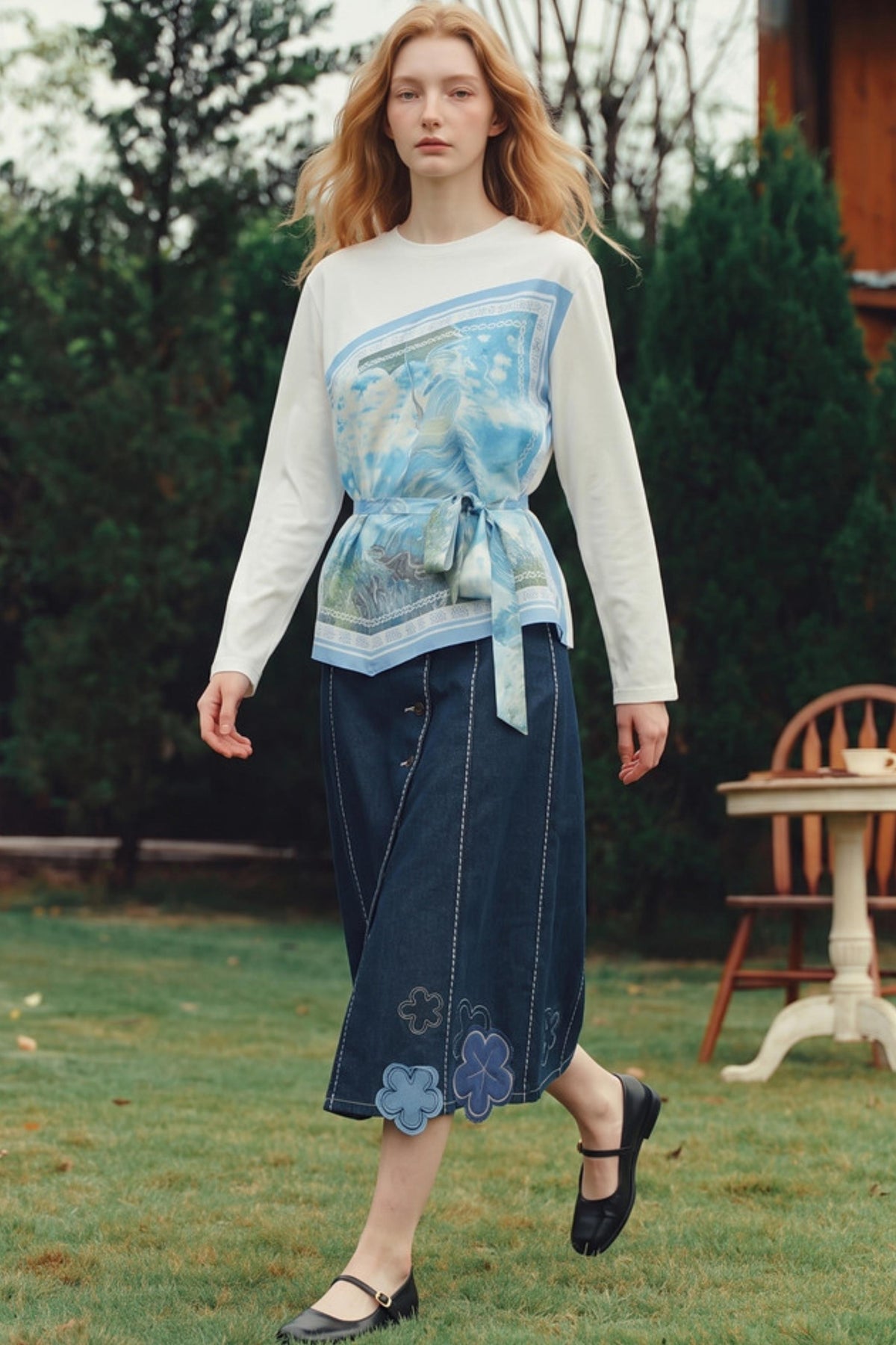 A woman stands outdoors on a grassy lawn wearing a white long-sleeve top with a blue patterned wrap-style panel and tie detail, paired with a dark blue midi skirt featuring blue floral embroidery near the hem, and black Mary Jane shoes.