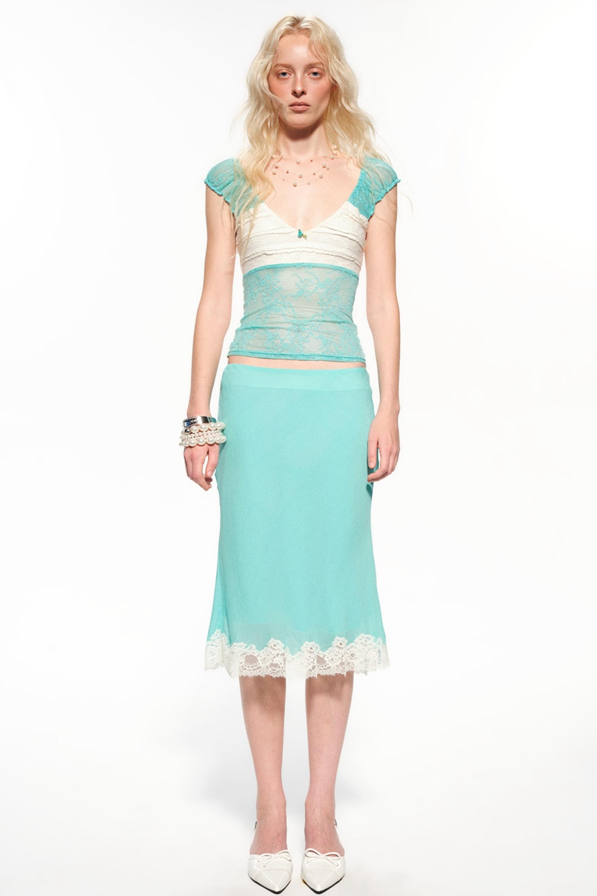 A blonde model wearing a two-piece outfit consisting of a light aqua blue midi skirt with white lace trim and a matching sheer lace crop top with white underlayer, photographed against a solid white studio background.