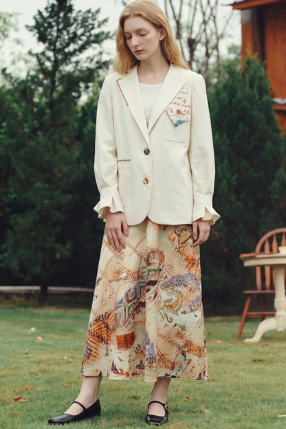 A woman stands outdoors on a grassy lawn wearing a cream-colored blazer with gold buttons and embroidery, paired with a long beige maxi skirt featuring a colorful vintage map print, and black Mary Jane shoes.