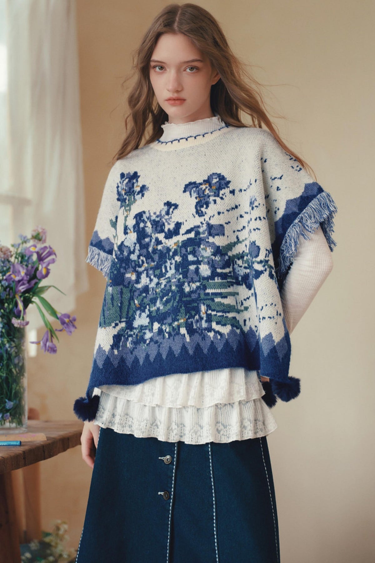 A woman wearing a cream and blue patterned knit poncho sweater layered over a white ruffled top and a dark blue skirt, standing indoors near a window and flowers.