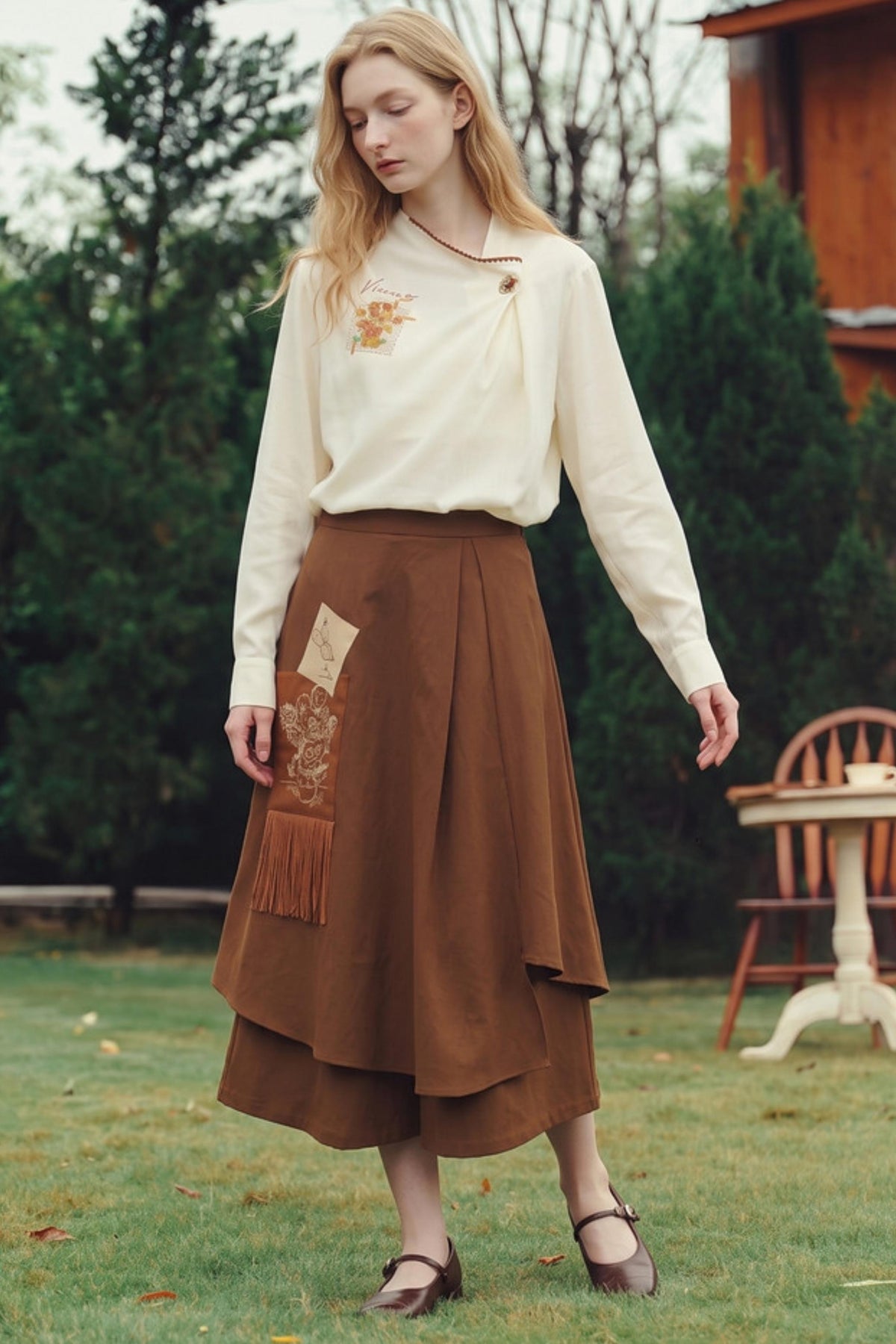 A woman stands outdoors on a grassy lawn wearing a cream long-sleeve blouse with a small floral embroidery on the chest and a brown tiered midi skirt with patchwork and fringe details. Trees and a wooden chair are visible in the background.