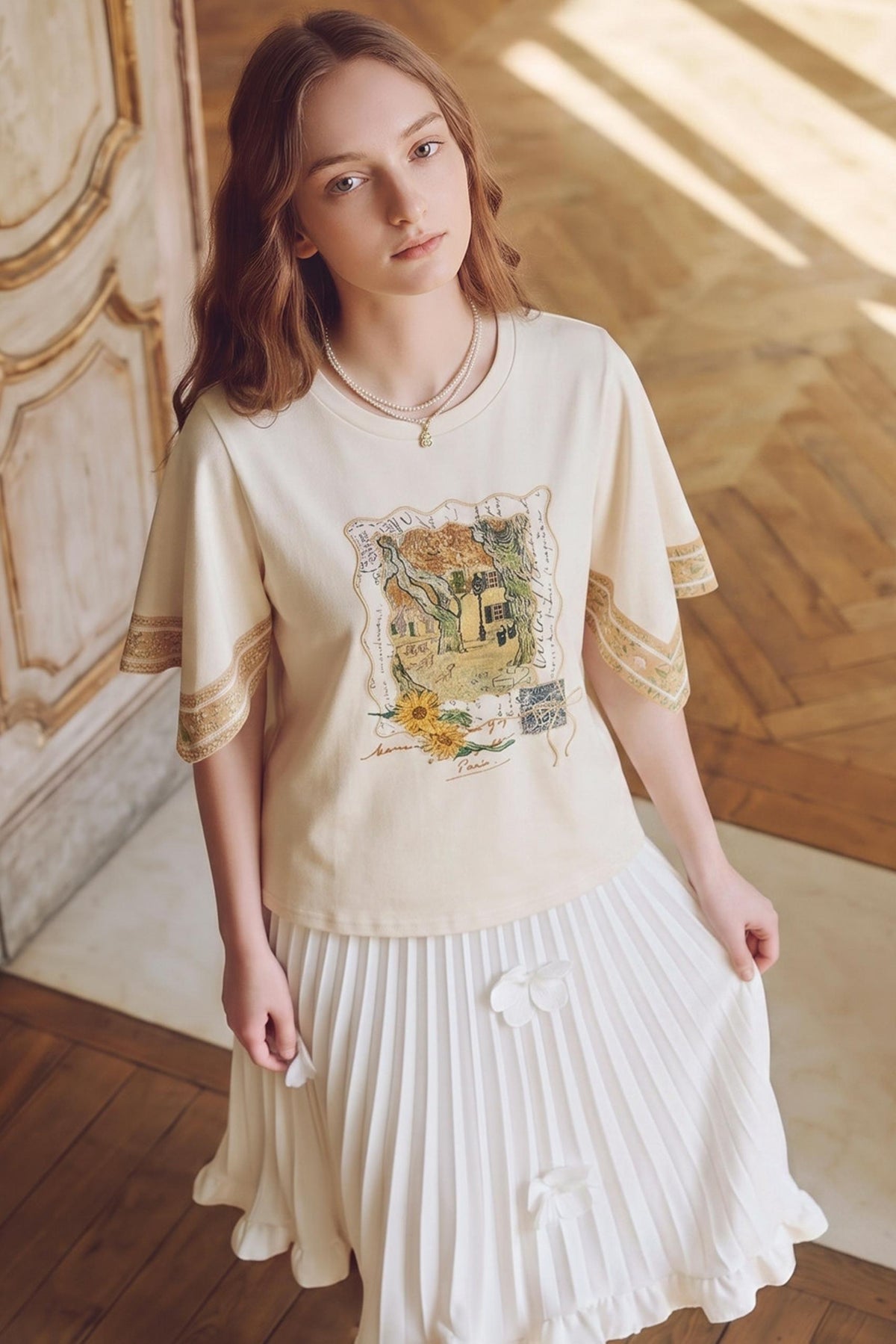 A young woman wearing a cream-colored short-sleeve t-shirt with a vintage-style botanical illustration on the front and gold-trimmed sleeve cuffs, paired with a white pleated skirt, standing in a sunlit room with wooden floors.