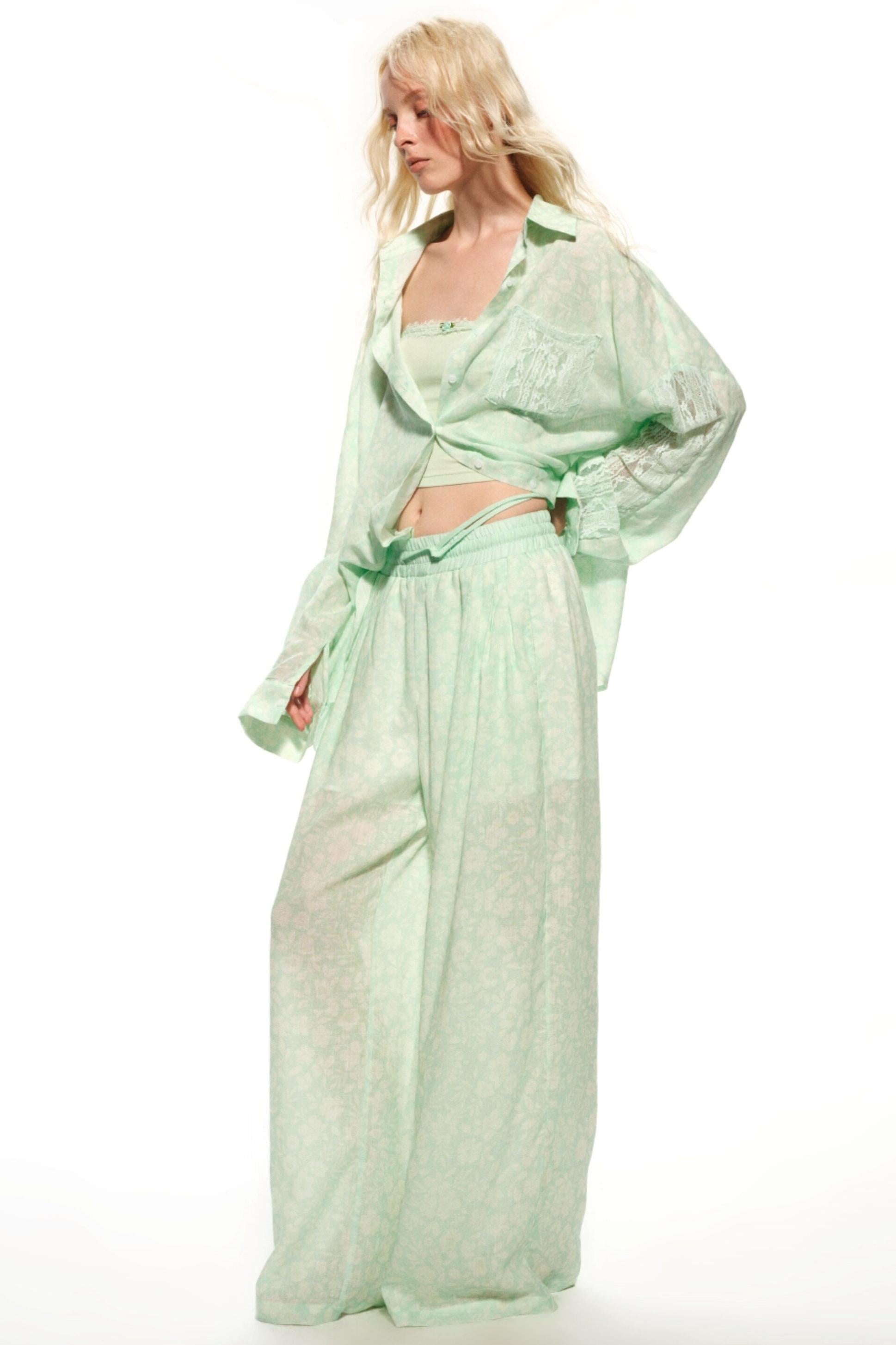 A blonde model wearing a light mint green two-piece outfit consisting of a long-sleeve button-up shirt tied at the waist and matching wide-leg pants, photographed against a solid white background.