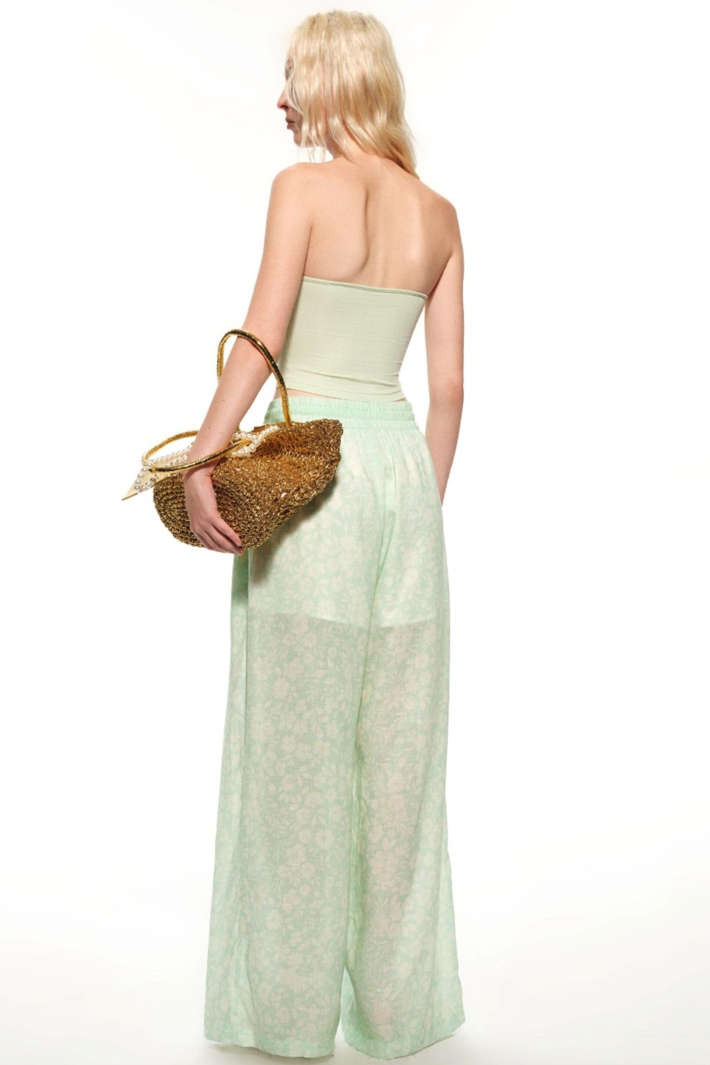 A woman shown from the back wearing a pale mint green strapless tube top and matching wide-leg sheer pants, holding a woven straw handbag against a plain white studio background.