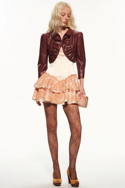 A model wearing a dark brown faux-leather cropped jacket with ruffles, a cream-colored top, a peach ruffled skirt, patterned tights, and brown platform heels, holding a small beige clutch against a white studio background.