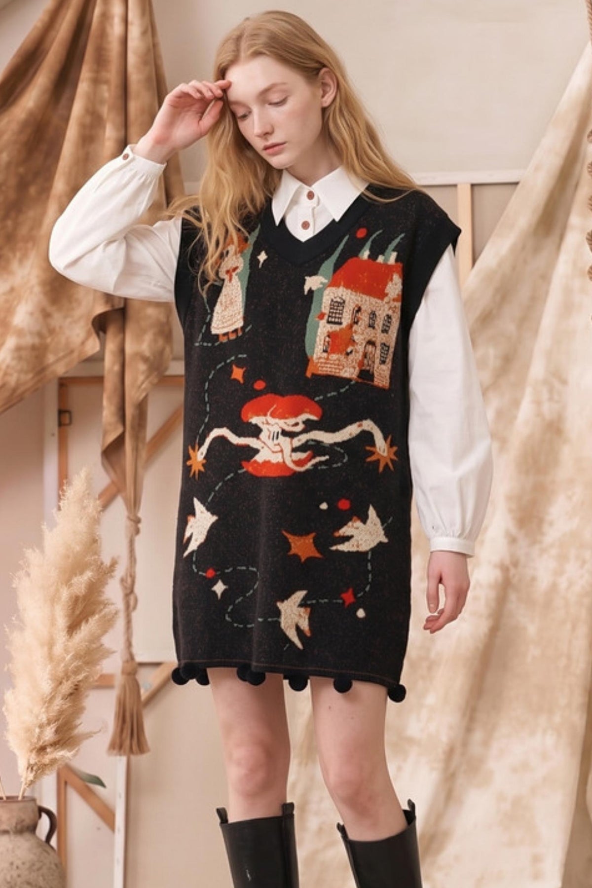 A woman modeling a black knit sweater vest with whimsical, colorful embroidered motifs including a skeleton, a house, stars, and birds, worn over a white collared shirt and black boots.