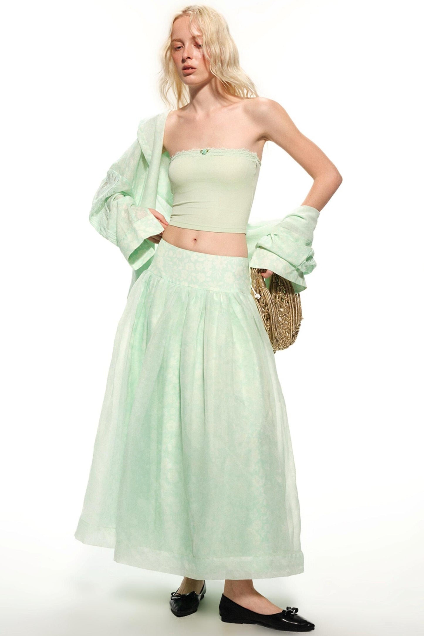 A model wearing a light mint green two-piece set consisting of a strapless tube top and a matching long pleated skirt, styled with a matching sheer jacket draped off one shoulder, black flats, and a woven clutch bag against a white background.