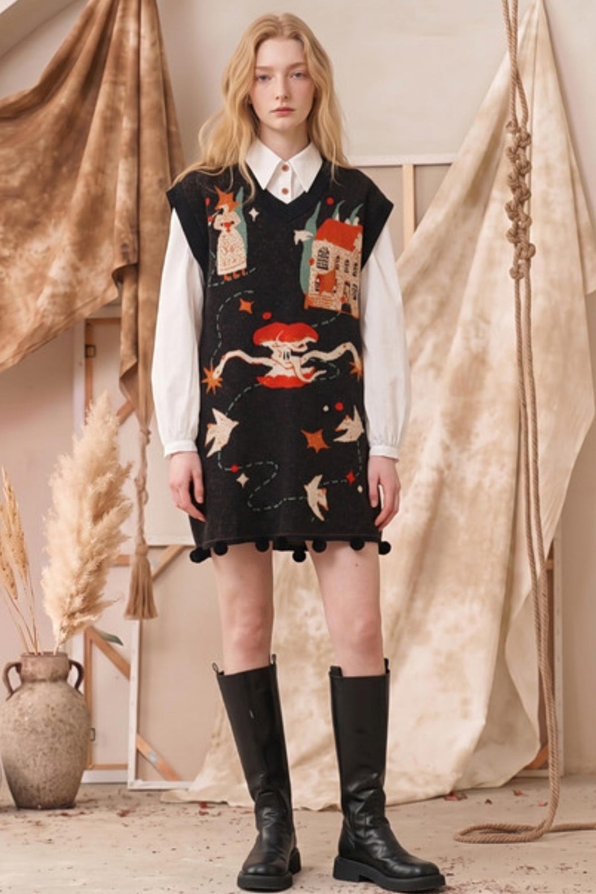 A model stands in a styled studio setting wearing a black knit sweater vest dress with whimsical embroidered motifs, layered over a white collared shirt and paired with black knee-high boots.