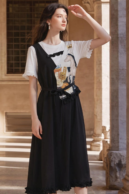 A woman models a black sleeveless pinafore-style dress over a white ruffled short-sleeve t-shirt with a graphic print, standing in a sunlit stone corridor.
