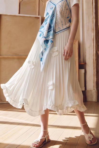 A woman wearing a flowing white pleated midi skirt with 3D floral appliques, a white top, and a blue patterned silk scarf, standing on a wooden floor.