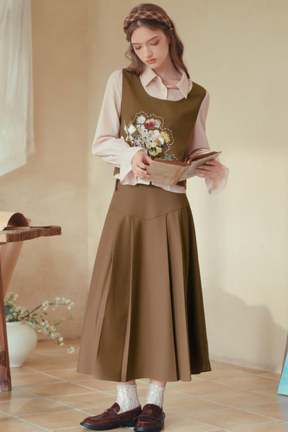 A woman stands indoors wearing a brown pleated midi skirt with a matching brown vest layered over a light pink blouse, holding a book. She is wearing brown loafers and white socks in a warm, neutral-toned room.