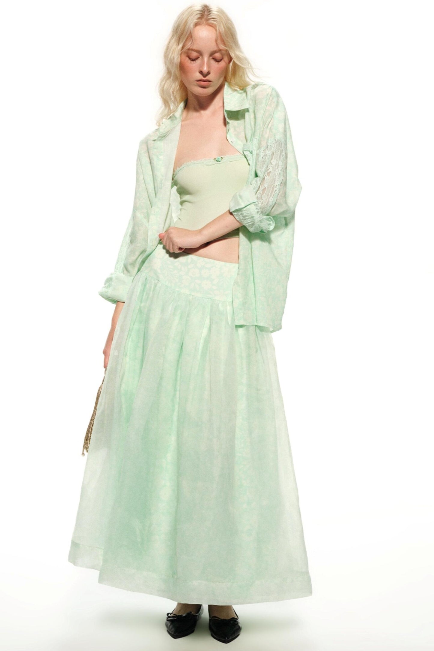 A blonde model wearing a light mint green two-piece outfit consisting of a strapless tube top and a matching long sheer lace-trimmed skirt, styled with an open long-sleeve button-up shirt and black flats against a white studio background.