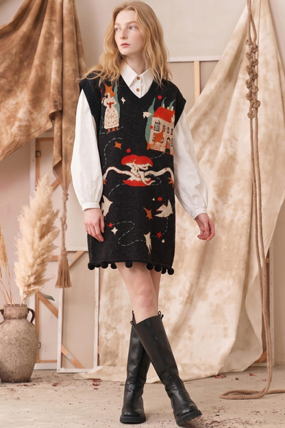 A model stands in a rustic studio setting wearing a black knit sweater vest with whimsical embroidered illustrations of houses, stars, and a cat, layered over a white collared shirt and paired with black leather boots.