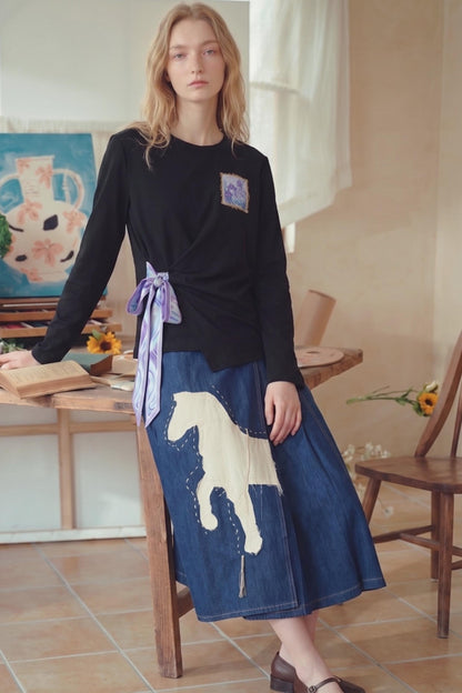 A woman stands indoors wearing a black long-sleeve wrap-style top with a small square patch on the chest and a blue denim skirt featuring a large cream horse graphic. She is posed beside a wooden table in a softly lit room with chairs and wall art visible.