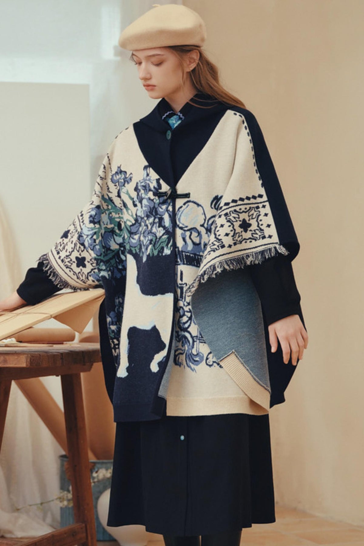A woman wearing a beige beret and a long-sleeved cape-style coat with a cream base, navy panels, and blue floral/bird motifs, standing indoors near a wooden table.