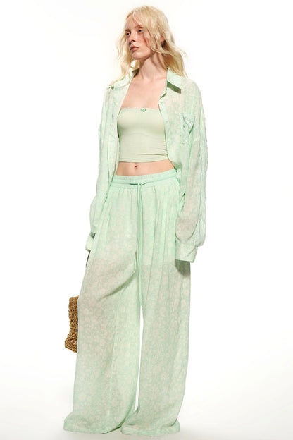 A blonde model wearing a light mint green two-piece lounge set consisting of a cropped tank top and matching wide-leg pants, styled with a sheer matching open-front shirt and a small woven bag against a white background.