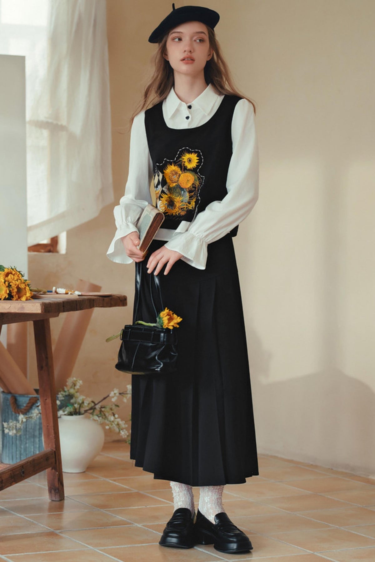 A woman stands indoors wearing a black sleeveless vest and long black skirt over a white long-sleeve collared blouse, with a black beret, white socks, black loafers, and a black handbag with yellow floral embroidery.