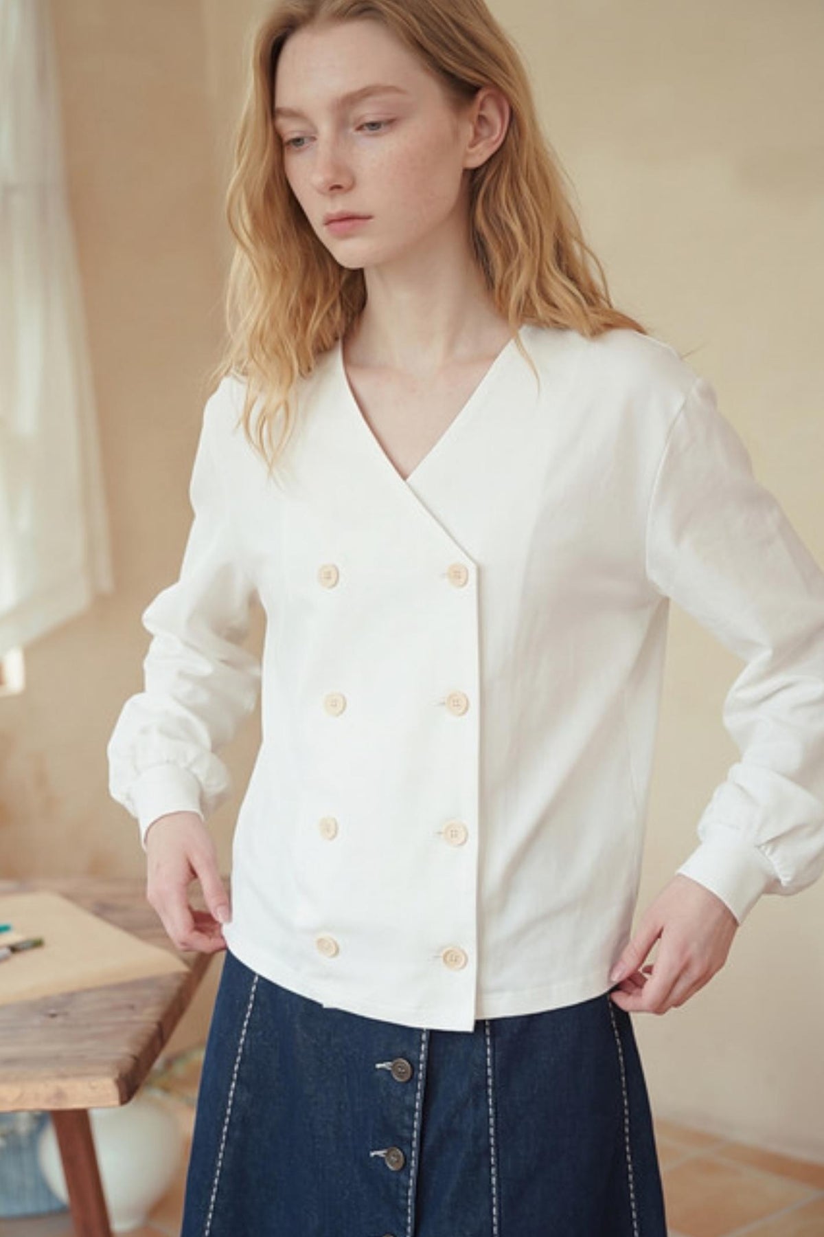 A woman wearing a white long-sleeve wrap-style blouse with decorative buttons, paired with a dark blue skirt, in a softly lit indoor setting.