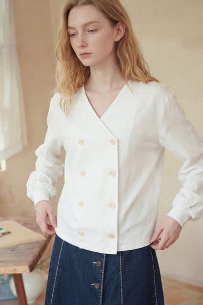 A woman wearing a white long-sleeve wrap-style blouse with decorative buttons, paired with a dark blue skirt, in a softly lit indoor setting.