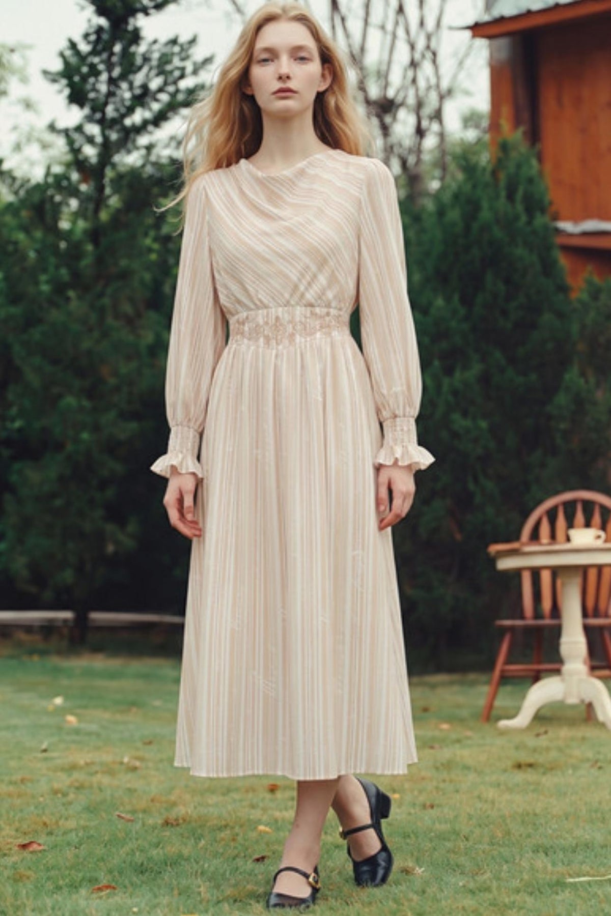 A woman stands outdoors in a grassy yard wearing a long-sleeve midi dress with vertical pleats and ruffled cuffs. The dress is a light warm cream or beige color.