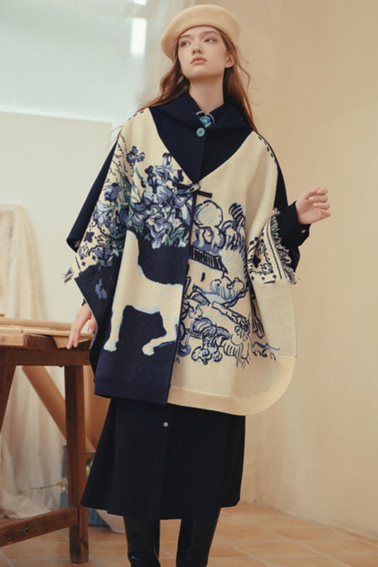 A model wearing a cream and navy blue patterned poncho over a black dress, paired with a beige beret, in an indoor setting.