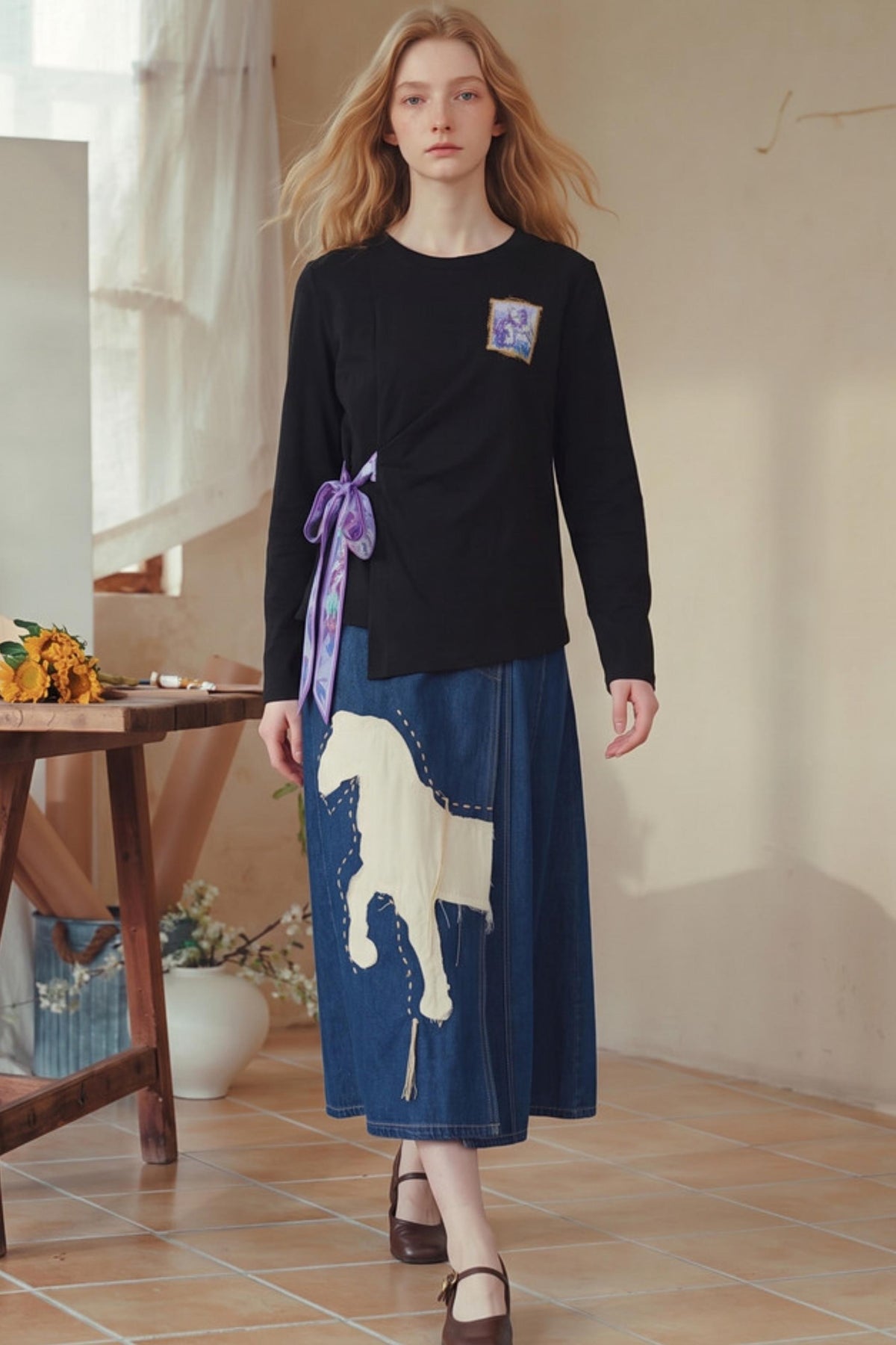 A woman models a black long-sleeve wrap top with a purple ribbon tie and a blue denim skirt featuring a cream-colored horse graphic, in a rustic indoor setting.