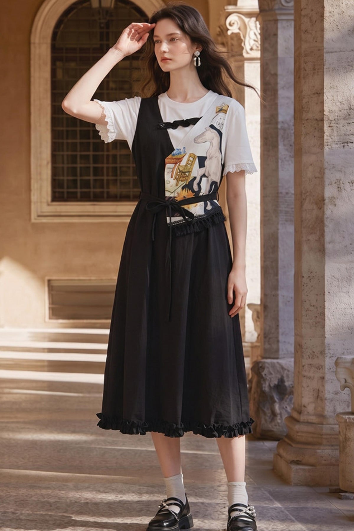 A woman stands in a sunlit architectural courtyard wearing a black sleeveless midi dress over a white graphic T-shirt, paired with white socks and black loafers.