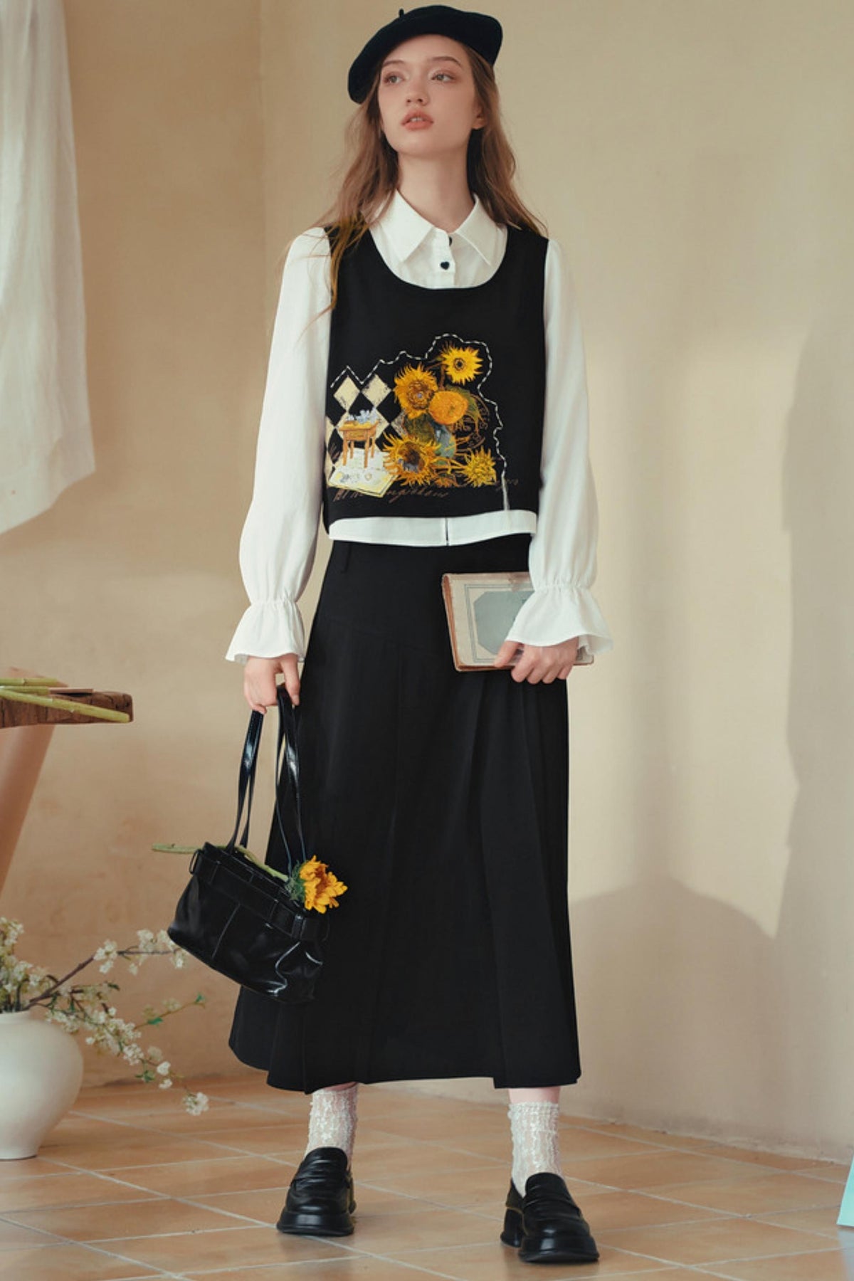 A model stands in a warm-toned indoor setting wearing a black sleeveless vest over a white long-sleeve collared blouse, paired with a long black skirt, black loafers, white socks, a black beret, and carrying a black handbag with a yellow flower accent.