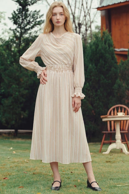 A woman stands outdoors on a grassy lawn wearing a long-sleeve midi dress with vertical stripes in beige and cream, featuring a ruffled waistband and black flat shoes.