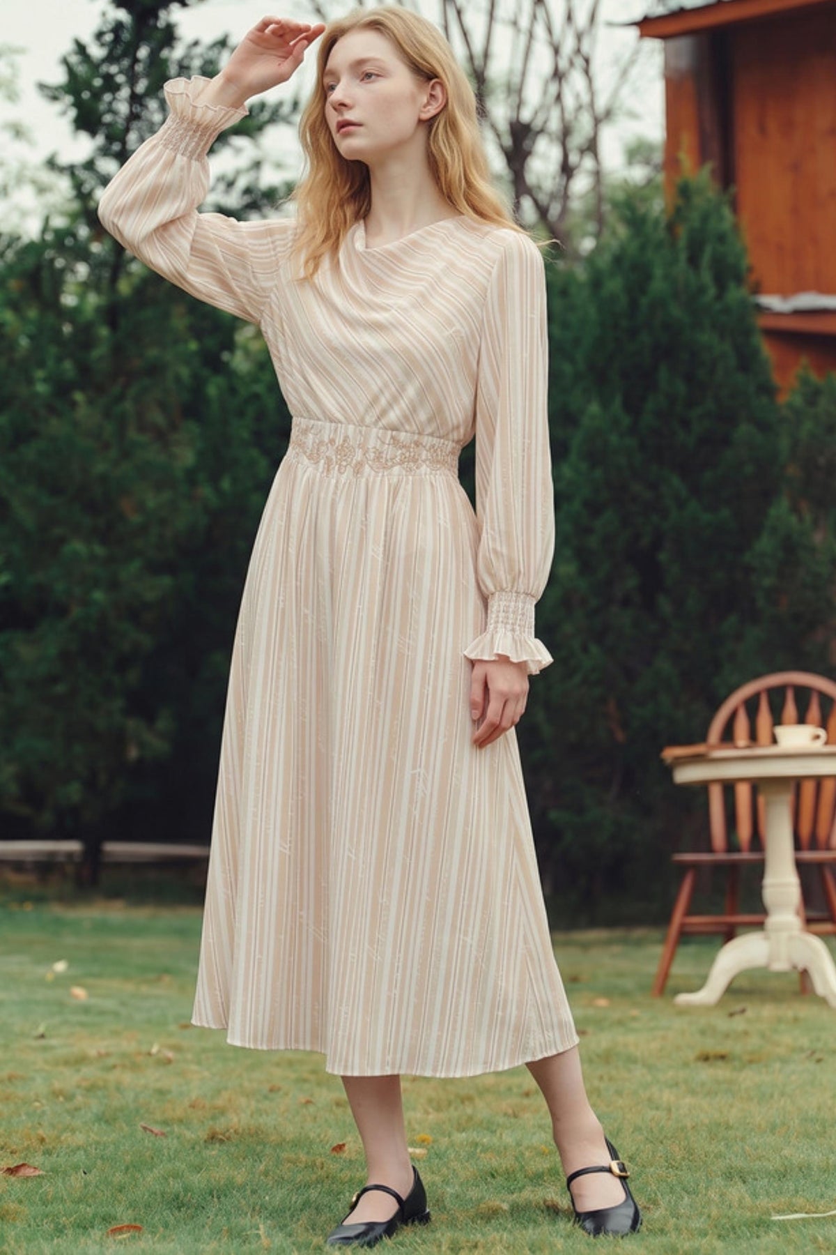 A woman stands outdoors on a grassy lawn wearing a long-sleeve midi dress with vertical stripes in cream and light tan. The dress features a gathered waist and ruffled cuffs.