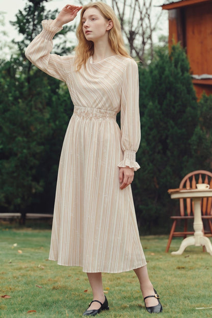 A woman stands outdoors on a grassy lawn wearing a long-sleeve midi dress with vertical stripes in cream and light tan. The dress features a gathered waist and ruffled cuffs.