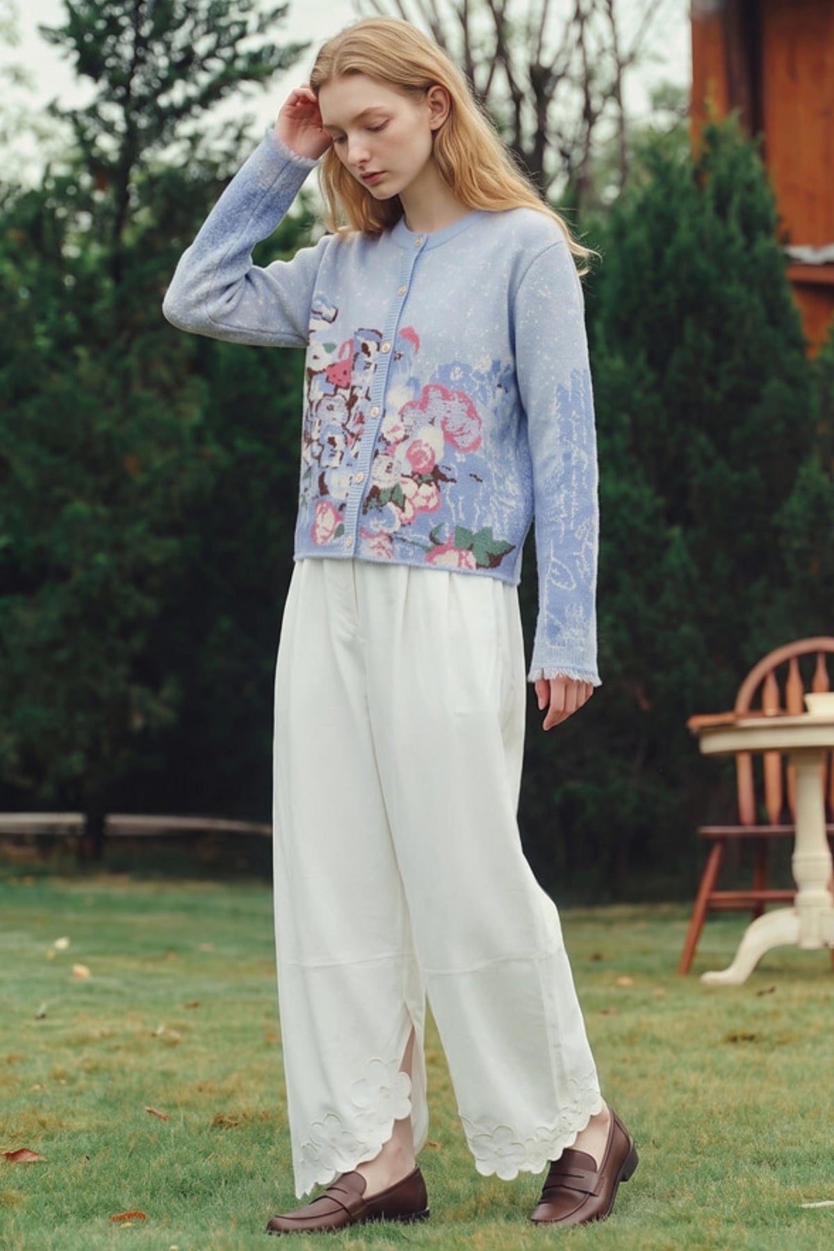 A woman standing outdoors in a grassy yard wearing a light blue cropped cardigan with a colorful floral print on the front, white wide-leg pants, and brown loafers.