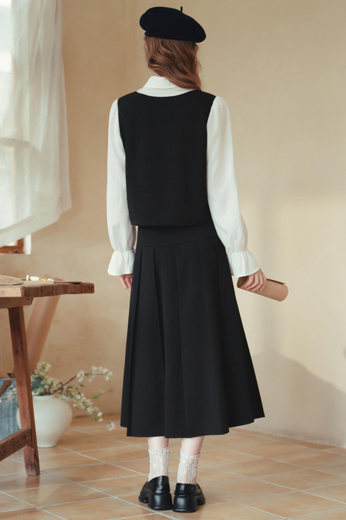 A woman is shown from behind wearing a black sleeveless vest and pleated midi skirt over a white long-sleeve blouse, paired with a black beret, white socks, and black loafers in a warm-toned indoor setting.