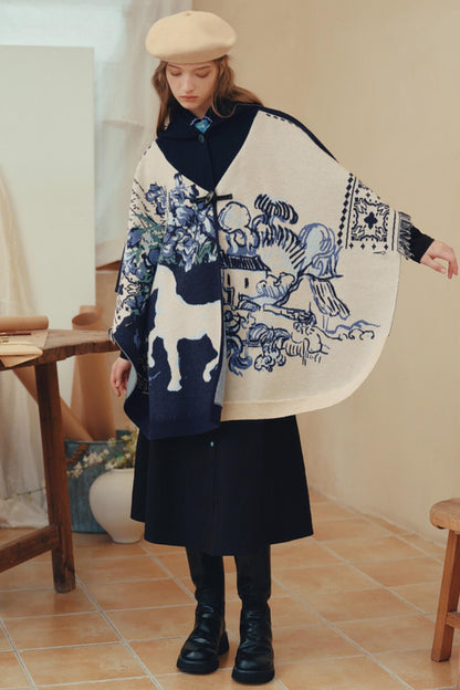 A model wearing a cream and navy blue poncho-style shawl with blue and white traditional-style illustrations, styled with a dark skirt, black boots, and a beige beret in a warm-toned indoor setting.