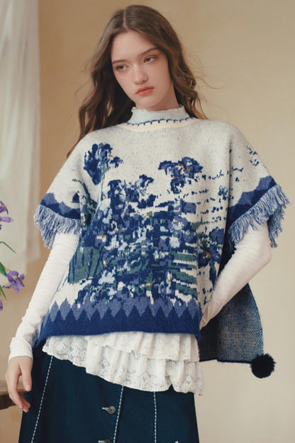A woman wearing a blue and white patterned knit poncho sweater over a white long-sleeve top and dark skirt, photographed indoors against a soft beige background.