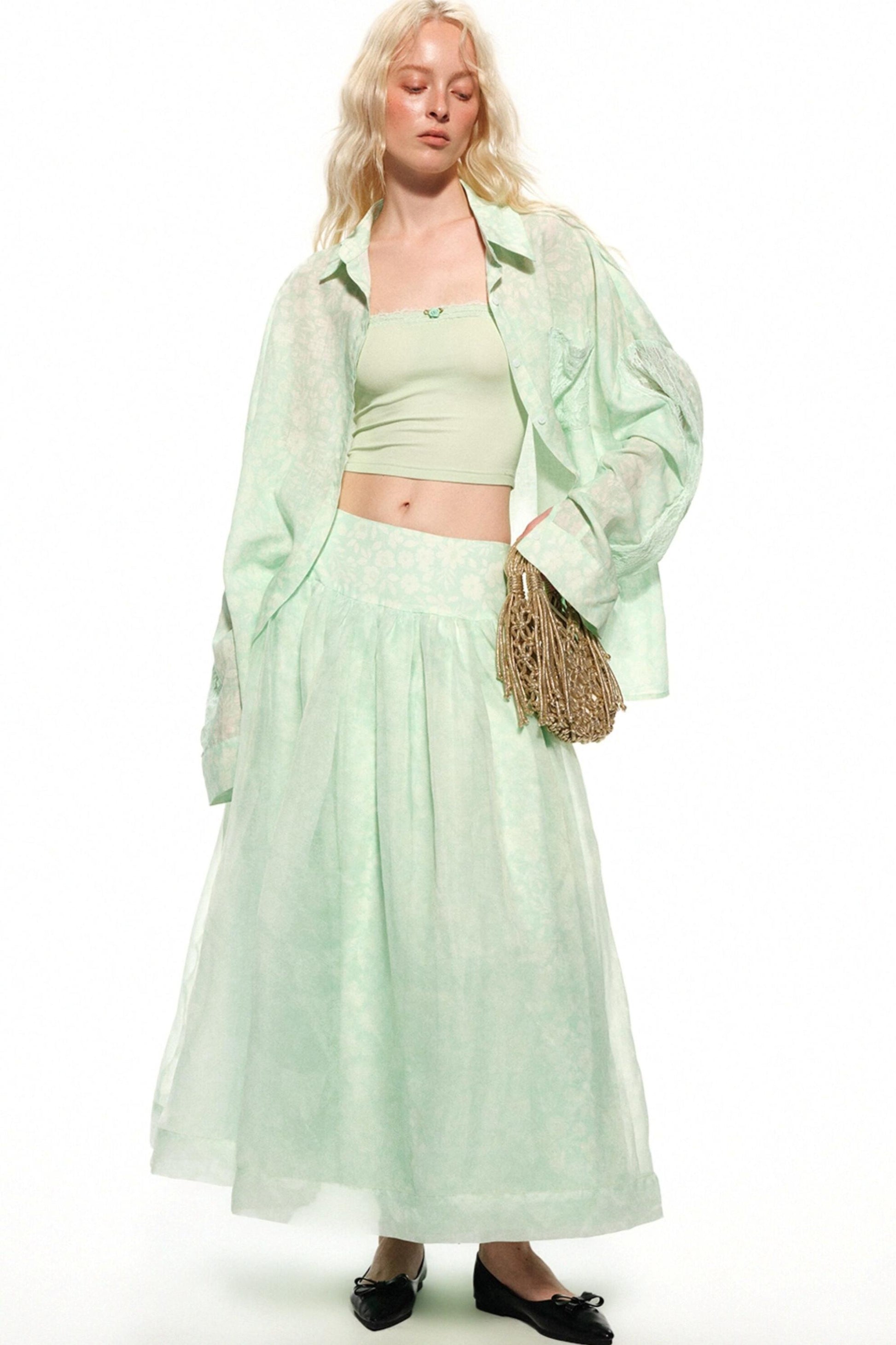 A model wearing a light mint green two-piece set consisting of a cropped tank top and a matching pleated maxi skirt, styled with an oversized sheer matching button-up shirt and a woven straw bag, on a white studio background.