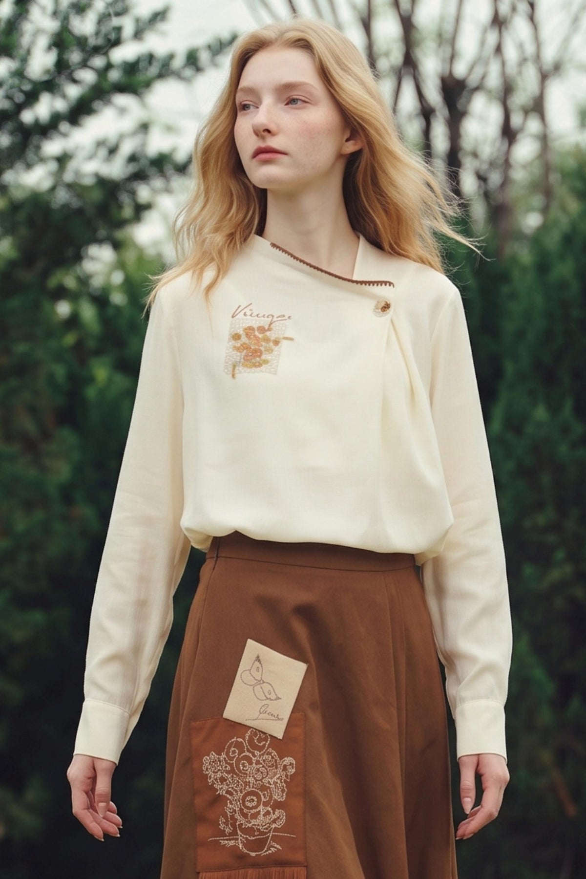 A woman standing outdoors in front of a blurred green forest background, wearing a cream-colored long-sleeve top with a small embroidered patch on the chest and a brown skirt with a decorative patch on the front pocket.