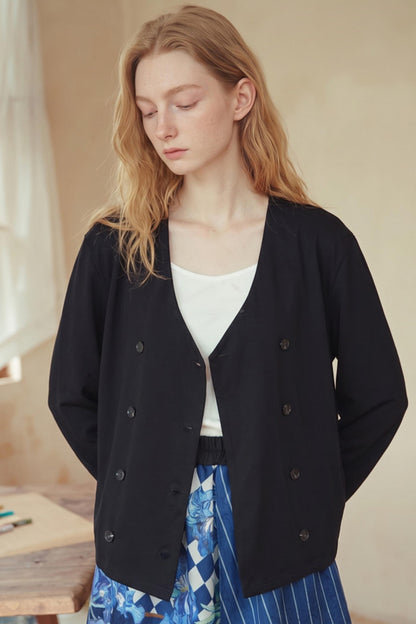 A woman wearing a black double-breasted cardigan over a white top and a blue patterned skirt, standing in a softly lit indoor setting.