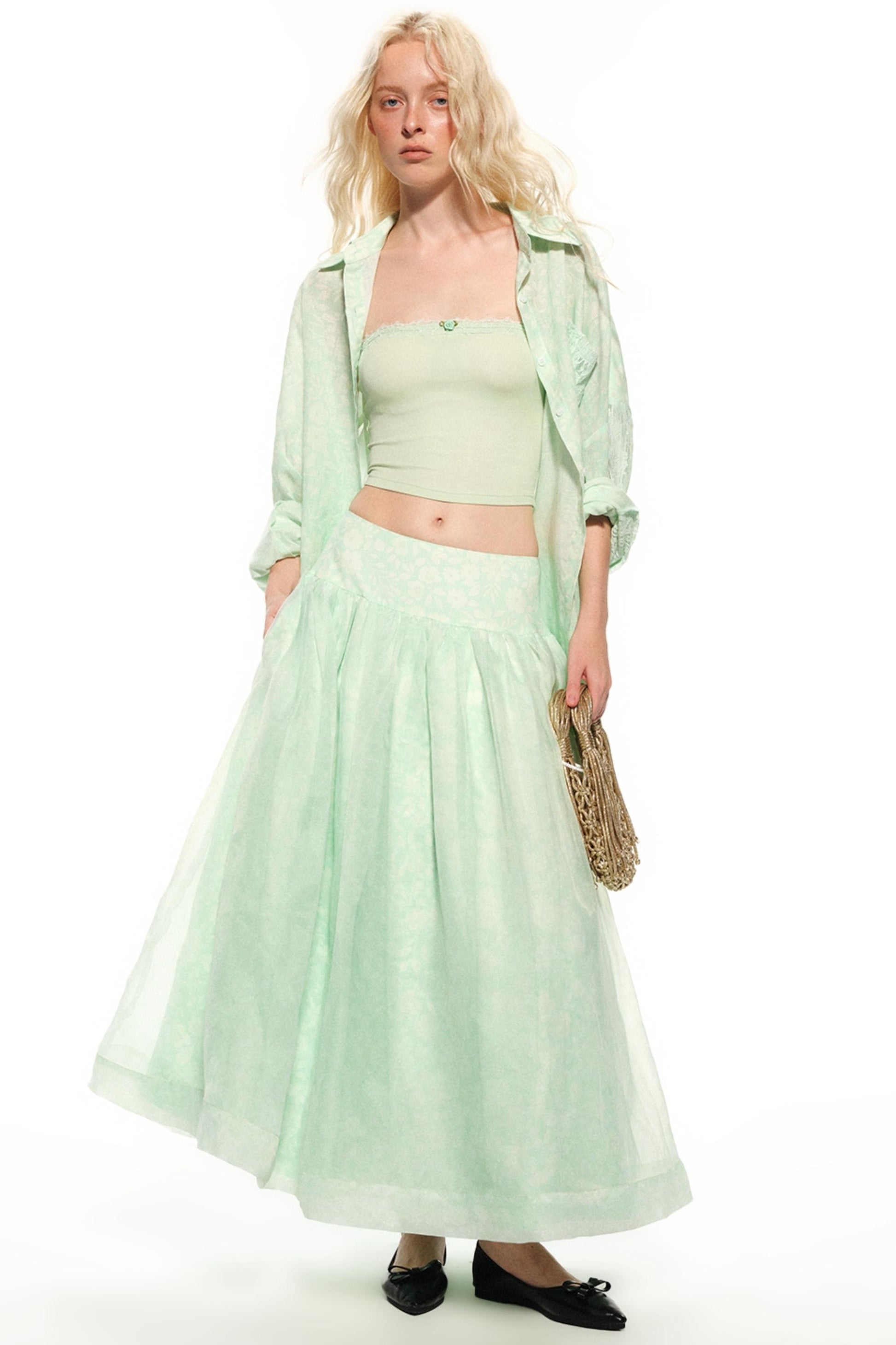 A blonde model wearing a light mint green three-piece outfit consisting of a cropped tube top, a matching long pleated skirt, and an open lightweight button-up shirt, holding a woven straw bag against a white studio background.