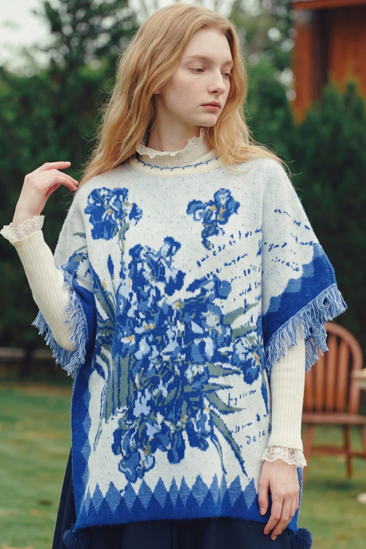 A woman wearing a blue and white knit poncho sweater with a floral pattern and fringe, photographed outdoors in a garden setting.