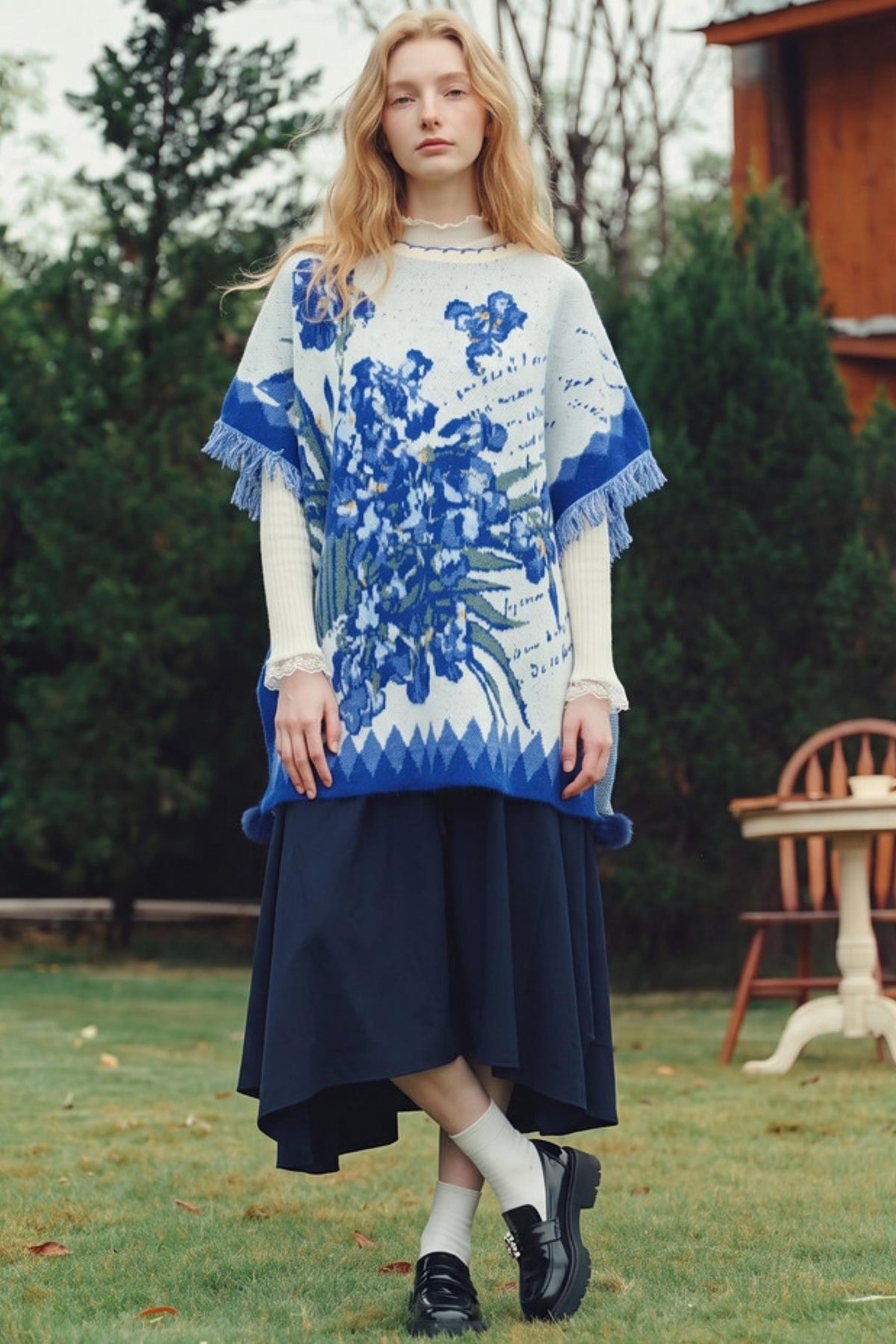A woman stands outdoors on a grassy lawn wearing a cream and blue floral knit poncho sweater over a dark navy midi skirt, with white socks and black loafers.