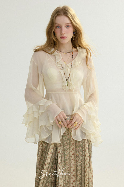 Woman wearing a sheer beige blouse with ruffled details and patterned pants on a white background.
